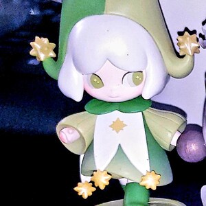 May include: A small figurine of a character wearing a two-toned green jester hat and a matching green and white outfit. The figure has white hair, light green eyes, and is decorated with several small yellow star-shaped accents on the hat and hem.