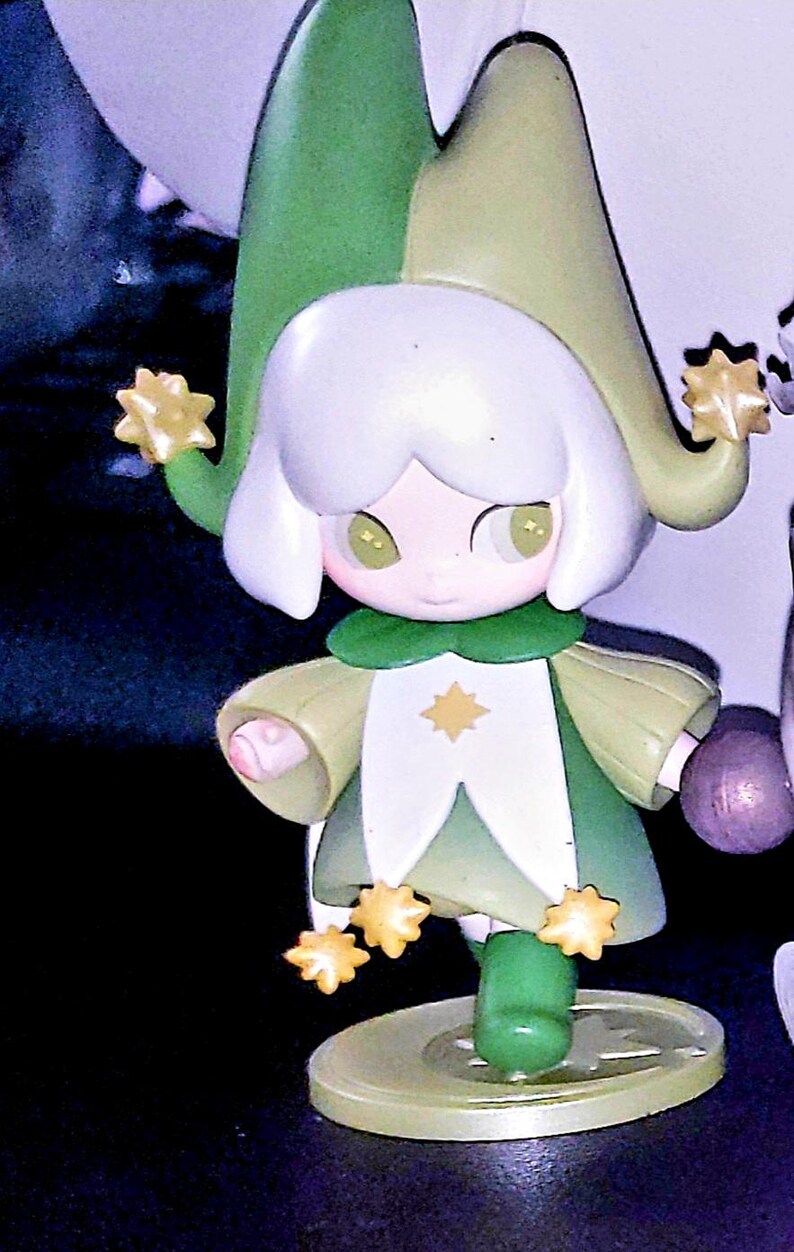 May include: A small figurine of a character wearing a two-toned green jester hat and a matching green and white outfit. The figure has white hair, light green eyes, and is decorated with several small yellow star-shaped accents on the hat and hem.