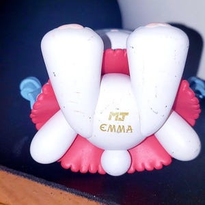 May include: A white figurine with pink accents, featuring two prominent white feet and a round tail. The text MJ EMMA is printed in gold on the central white section. The figure has light blue appendages extending from the sides and a pink ruffled base.