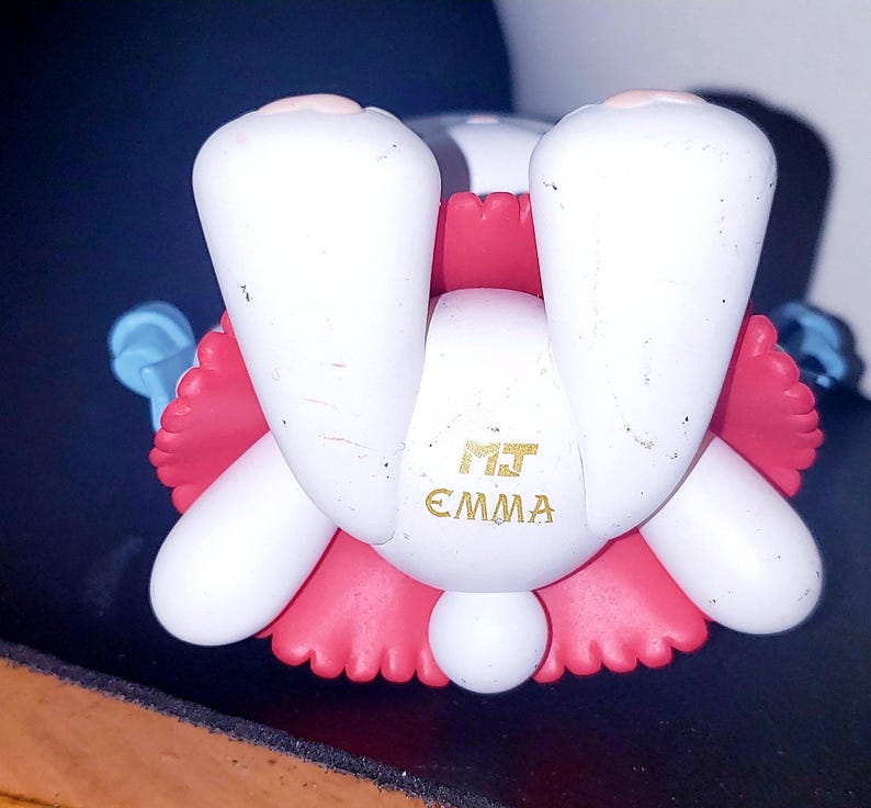May include: A white figurine with pink accents, featuring two prominent white feet and a round tail. The text MJ EMMA is printed in gold on the central white section. The figure has light blue appendages extending from the sides and a pink ruffled base.