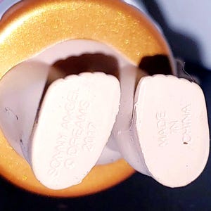May include: The underside of a figurine's feet, featuring light-colored skin tones. The left foot is embossed with the text SONNY ANGEL, &copy; DREAMS, 2017, and the right foot is embossed with the text MADE IN CHINA. A metallic gold circular base surrounds the feet.
