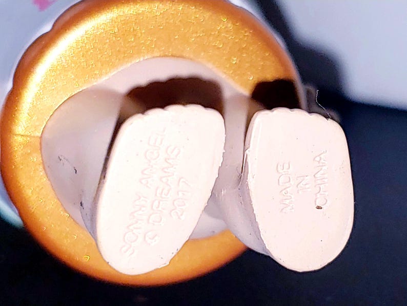 May include: The underside of a figurine's feet, featuring light-colored skin tones. The left foot is embossed with the text SONNY ANGEL, &copy; DREAMS, 2017, and the right foot is embossed with the text MADE IN CHINA. A metallic gold circular base surrounds the feet.