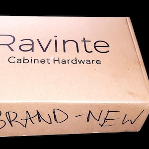 May include: A cardboard box with the text "Ravinte Cabinet Hardware" printed on the top. The words "BRAND-NEW" are handwritten on the front of the box. The box is tan.