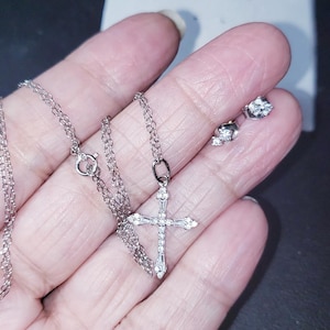 May include: A silver necklace with a cross pendant and matching stud earrings. The cross pendant and earrings are adorned with small, sparkling gemstones. The necklace chain is delicate and the jewelry is displayed on a light background.