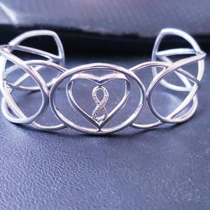 May include: A silver-colored cuff bracelet with an open design. The bracelet features a heart-shaped center with an infinity symbol and small stones. The sides have an intricate, interwoven pattern.