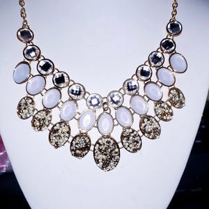 May include: A gold-tone statement necklace featuring a multi-layered bib design. The piece is adorned with alternating oval-shaped white cabochons, clear faceted crystals, and textured gold-tone stones with a sparkling, granular surface finish.