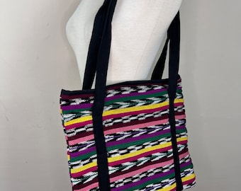 May include: A colourful striped tote bag with black straps. The bag is made of woven fabric and has a variety of colours, including pink, green, yellow, and black.