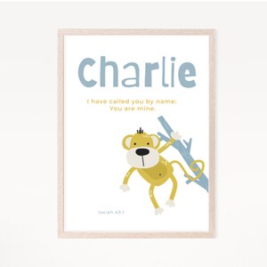May include: A framed print with a light blue background and a yellow monkey holding a branch. The print has the name "Charlie" and the text "I have called you by name: You are mine. Isaiah 43:1".