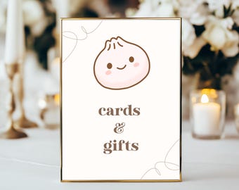 May include: A gold-framed sign with a white background and a cartoon illustration of a pink dumpling with a smiling face. The text "cards & gifts" is written in brown letters below the dumpling.