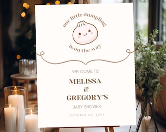 May include: A cream-coloured baby shower welcome sign on an easel. The sign features a cartoon dumpling illustration and the text "our little dumpling is on the way." It welcomes guests to Melissa & Gregory's baby shower, scheduled for October 25, 20XX.