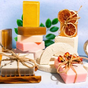 May include: An assortment of handmade soaps in various shapes and colors, including yellow, brown, pink, and white. Some soaps are decorated with dried orange slices, flowers, and twine. A shaving soap with a label is also visible.