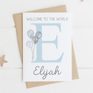 May include: A baby announcement card with the text "WELCOME TO THE WORLD" and the name "Elijah". The card features a large blue letter "E" and a cluster of gray and silver balloons. It is placed on a brown envelope, with wooden star decorations.