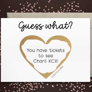 May include: A white card with the text "Guess what?" and "You have tickets to see Charli XCX!" inside a gold heart. The card has a "Scratch here" prompt and is on a dark brown surface with scattered glitter.