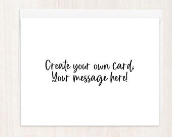 May include: A white greeting card with the text "Create your own card. Your message here!" in black script. The card is blank, offering space for a personalized message. The background is a light wood grain.