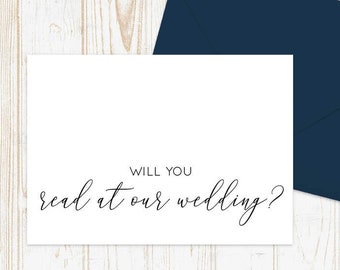 May include: A white card with black text that reads "Will you read at our wedding?" The card is on a white wooden background with a dark blue envelope.