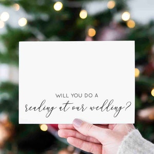 May include: A white card with black text that reads "Will you do a reading at our wedding?" The card is being held in front of a blurred background of a Christmas tree with lights.