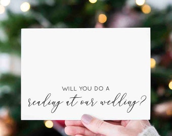 May include: A white card with black text that reads "Will you do a reading at our wedding?" The card is being held in front of a blurred background of a Christmas tree with lights.