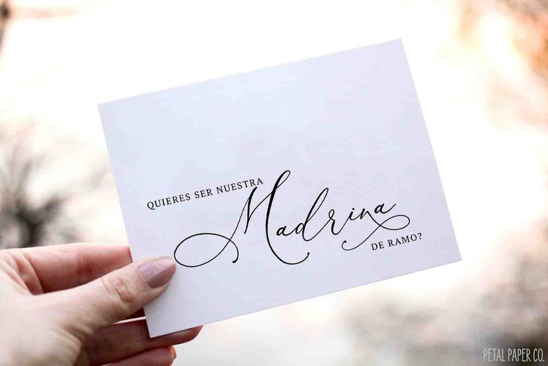 May include: A white card with black calligraphy text that reads "QUIERES SER NUESTRA Madrina DE RAMO?" The card is held by a person. The bottom right corner of the card has the text "PETAL PAPER CO."