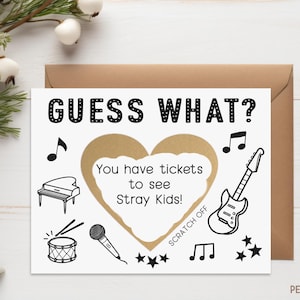 May include: A white card with black text that reads "GUESS WHAT?" and a gold heart-shaped scratch-off area that says "You have tickets to see Stray Kids!"  There are musical notes, a piano, a microphone, a drum, and a guitar drawn on the card.