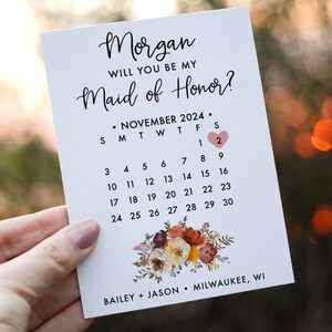 May include: A white card with the text "Morgan, Will you be my Maid of Honor?" and a calendar with a heart marking November 2nd, 2024. The card has a floral design at the bottom with the text "Bailey + Jason, Milwaukee, WI, Petal Paper Co."