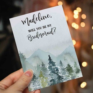 May include: A watercolor card with a mountain scene in shades of blue and green. The card says "Madeline, Will you be my Bridesmaid?"