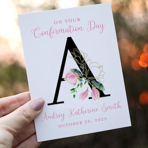 May include: A white card with pink and gold floral accents. The card says "On Your Confirmation Day" and features a large black letter "A" with pink flowers and gold accents. The name "Audrey Katherine Smith" and the date "October 20, 2025" are printed below the letter.