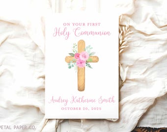 May include: A white card with a pink floral cross design. The text "ON YOUR FIRST Holy Communion" is at the top of the card. The name "Audrey Katherine Smith" and the date "OCTOBER 20, 2025" are at the bottom of the card.