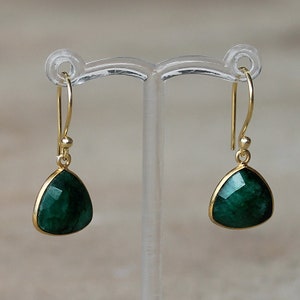 May include: A pair of gold dangle earrings with a green gemstone in the shape of a triangle. The earrings are on a clear display stand.