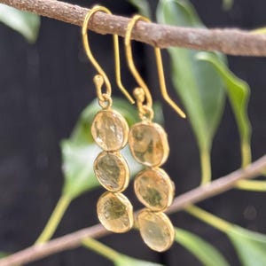May include: A pair of gold drop earrings featuring three vertically aligned, translucent, pale yellow circular stones. Each earring is suspended from a simple gold hook wire, showcasing a polished metal setting that frames the round, faceted gems.