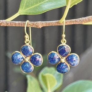 May include: A pair of gold-colored earrings, each featuring a four-petal flower design. The petals are a deep blue with hints of purple, set in a gold frame. The earrings hang from delicate gold hooks.