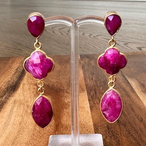 Peut inclure: Paire de boucles d'oreilles pendantes dorées. Chaque boucle d'oreille comporte trois pierres précieuses rose foncé facettées : une goutte d'eau en haut, une forme de trèfle à quatre feuilles au milieu et un ovale en bas. Les boucles sont présentées sur un présentoir transparent.