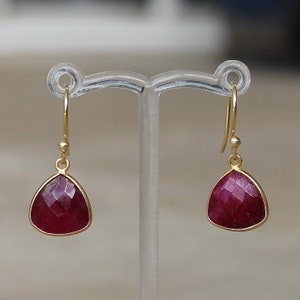 May include: A pair of gold earrings with a red gemstone. The earrings are teardrop shaped and have a simple design.
