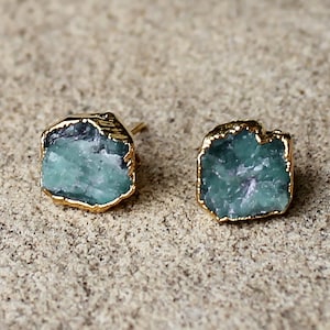 Puede incluir: Un par de pendientes de botón de piedra preciosa verde esmeralda con bordes dorados. Los pendientes tienen un aspecto crudo y natural, mostrando la textura y las variaciones de color únicas de las piedras. Los pendientes están sobre un fondo neutro.