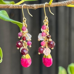 May include: Dangling earrings with gold-coloured hooks and chains. The earrings feature clusters of pink, magenta, and clear gemstones, culminating in a large, teardrop-shaped magenta stone at the bottom. The earrings are hanging from a brown branch.