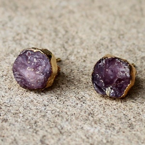 Könnte beinhalten: Ein Paar Ohrstecker mit grob geschnittenen, lila Edelsteinen und goldfarbenen Metallrändern. Die Ohrringe haben ein natürliches, organisches Aussehen und zeigen die rohe Schönheit der Steine. Die Ohrstecker sind vor einem neutralen Hintergrund platziert.