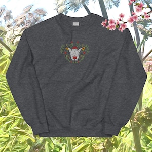 Könnte beinhalten: Ein dunkelgrauer Pullover mit einem weißen gestickten Design einer lächelnden, runden Figur mit roten Akzenten, umgeben von Blumen. Die Figur trägt einen roten Hut und ein rot-weißes Kleidungsstück.