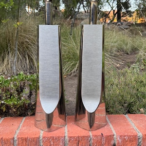 May include: Two polished metal table lamps with a brushed silver panel. The lamps have a sleek, modern design with a bulb at the top. The lamps are set against a backdrop of greenery and a red brick wall.