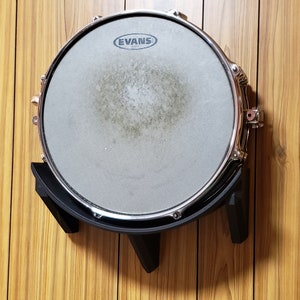 May include: A black drum rack with a silver snare drum mounted on it. The snare drum has an Evans logo on the head.