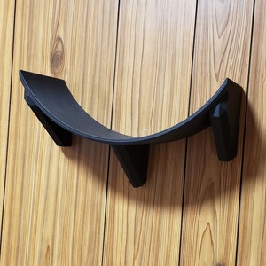 May include: A black plastic shelf bracket with three mounting points. The bracket is curved and designed to hold a round object.