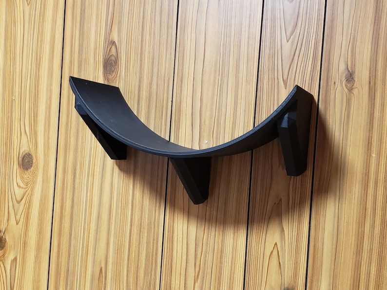 May include: A black plastic shelf bracket with three mounting points. The bracket is curved and designed to hold a round object.