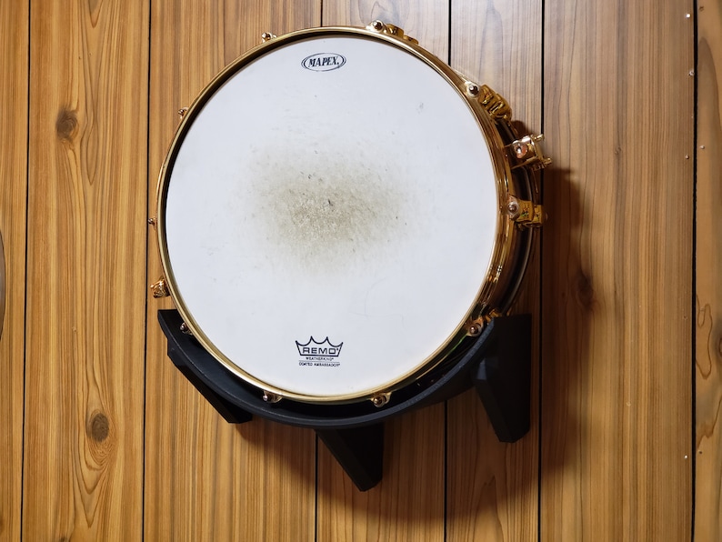 May include: A gold snare drum with a white Remo drumhead, mounted on a black wall mount. The drumhead has the text "MAPEX" and a Remo logo.