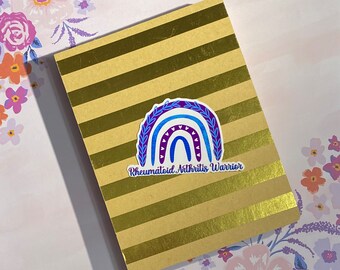 May include: A gold notebook with a blue and purple rainbow sticker that says "Rheumatoid Arthritis Warrior".