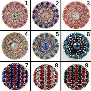 May include: Nine round, decorative snap buttons with various jewel-toned crystal arrangements. Each button features a central crystal surrounded by smaller gems in colors like pink, blue, teal, and red. The buttons are set in a metallic base.