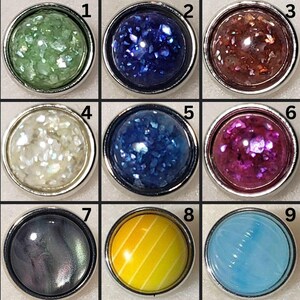 May include: Nine round decorative buttons with silver rims. The buttons feature various colors and textures, including green, blue, brown, white, purple, black, yellow, and light blue. Some buttons have a glittery appearance, while others have a smooth or patterned surface.