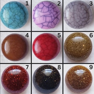 May include: Nine round, decorative cabochons in various colors and textures. The cabochons include shades of turquoise, purple, gray, brown, red, gold, and black. Some have a cracked or speckled appearance, while others are solid.