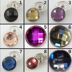 May include: Nine faceted snap buttons in various colors, including black, gold, purple, peach, fuchsia, and blue. Each button has a silver-colored base and a round, jewel-like top. The buttons are arranged in a grid.