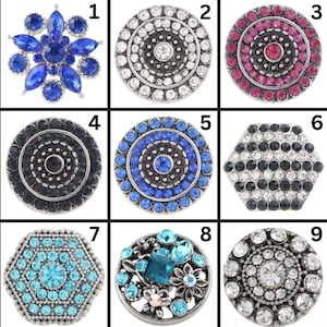 May include: Nine decorative snap jewelry buttons in various designs. The buttons feature a variety of colors, including blue, pink, black, and clear crystals. Some have floral or geometric patterns, while others have concentric circles.