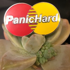 K&ouml;nnte beinhalten: Eine rote und gelbe Emaille-Anstecknadel mit dem Text "PanicHard" in silbernen Buchstaben.