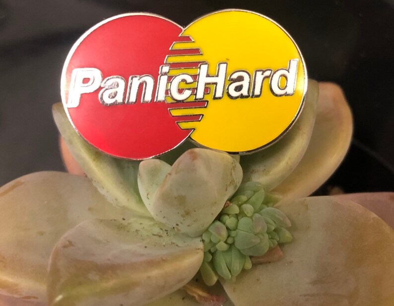 K&ouml;nnte beinhalten: Eine rote und gelbe Emaille-Anstecknadel mit dem Text "PanicHard" in silbernen Buchstaben.