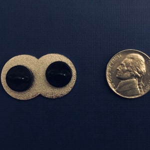 K&ouml;nnte beinhalten: R&uuml;ckansicht eines goldfarbenen Doppelkreis-Pins mit schwarzen Mittelpunkten. Der Pin ist etwa so gro&szlig; wie eine deutsche F&uuml;nf-Cent-M&uuml;nze.